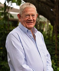 Smiling man with grey hair in light blue long sleeve shirt in a background of greenery