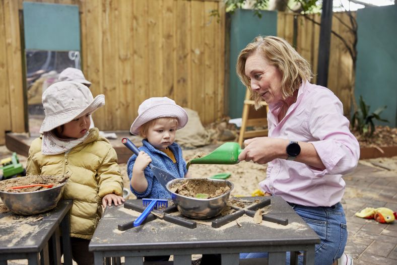 An elder woman playing with two younger children in a sandpit
