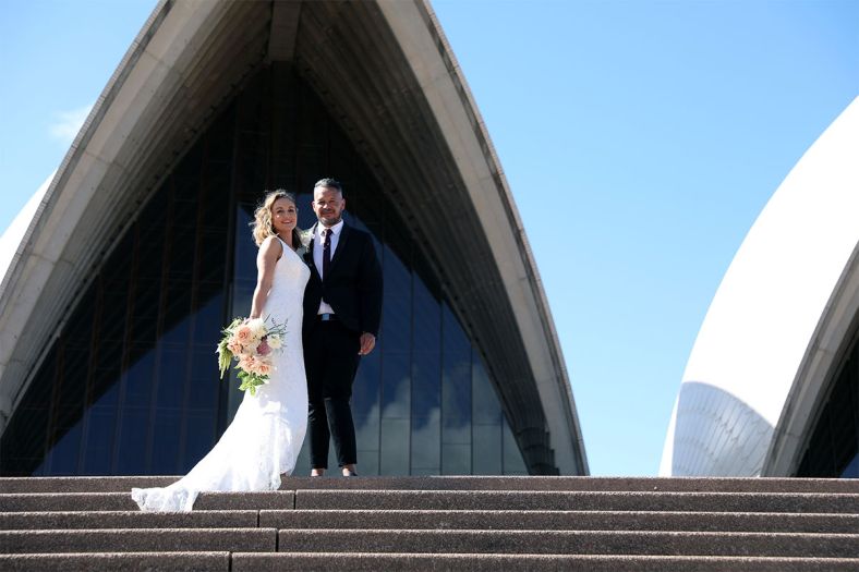 A bride and groom pose at the top of the Sydney Opera House steps