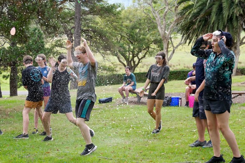 a group of eight young people wearing protective glasses, shorts and t-shirts stand on a large grassy area. there are trees in the background. Out of fram, someone is throwing water balloons which are bursting and spraying the group