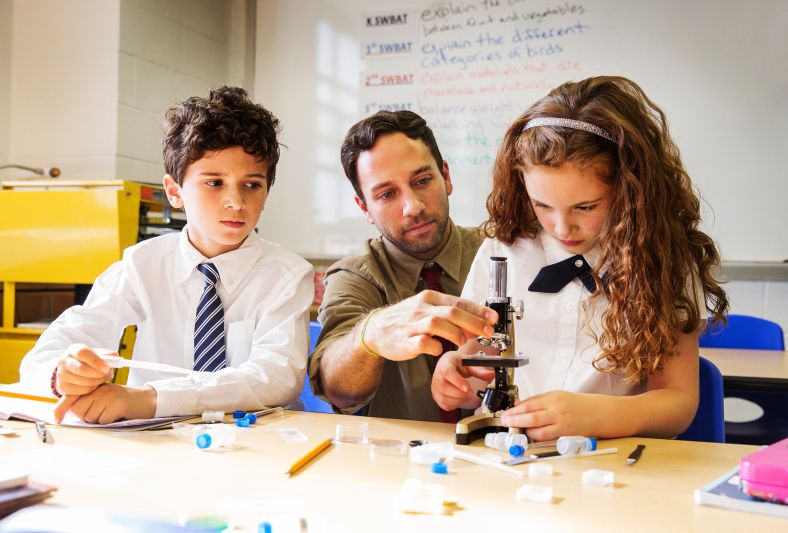 A teacher and two kids in a classroom using a microscope