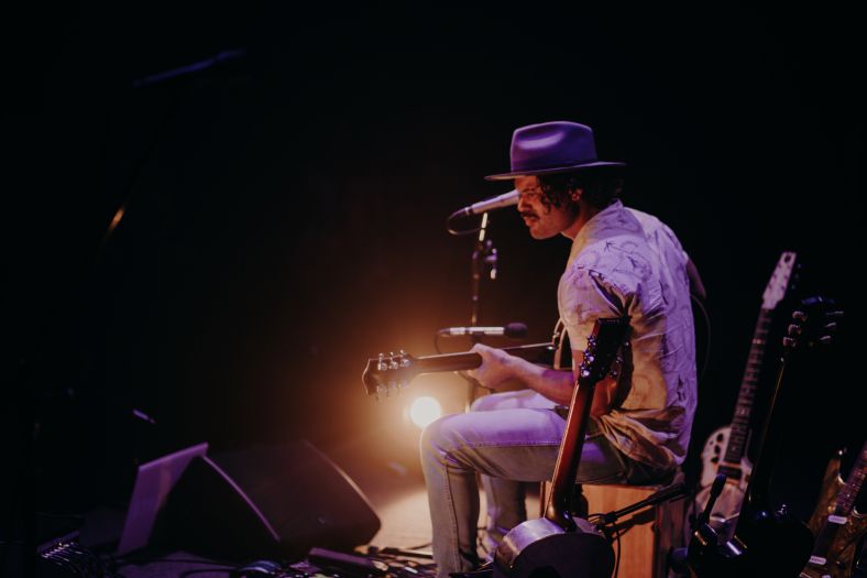 A man is sitting down facing away from the camera playing guitar. He is wearing a hat.