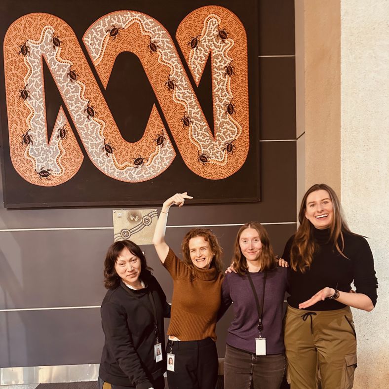 Four women stand together smiling under a painting of the ABC logo