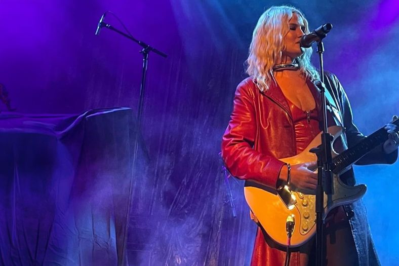 A woman is singing into a microphone. She is wearing red and holding a guitar.