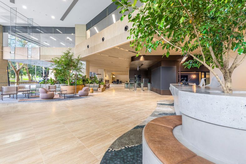 Foyer in office building with lounge chairs, tables, barrier entry to lifts for swipe cards, reception desk and windows looking into park.