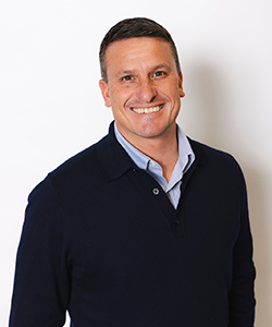 Smiling man with dark hair and dark blue jumper over a light blue shirt