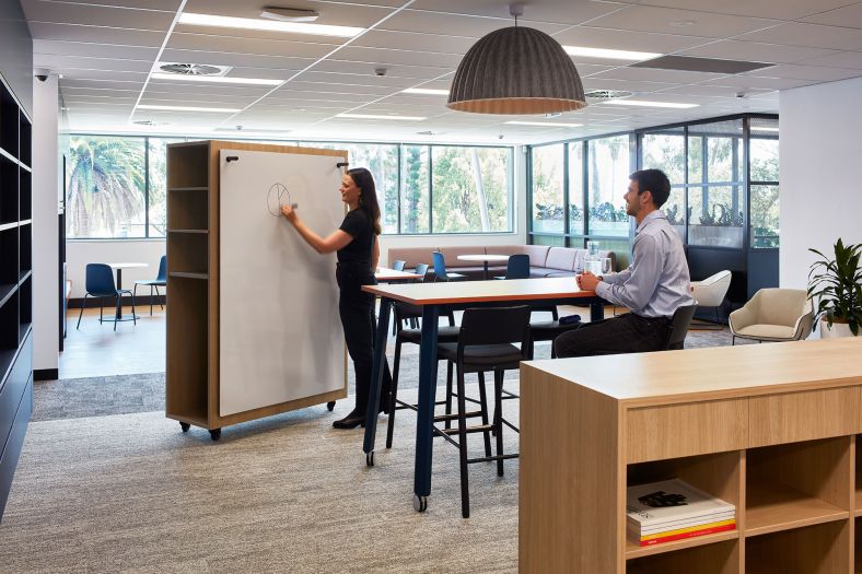 Woman writing on whiteboard while man sitting on stool watches the presentation.