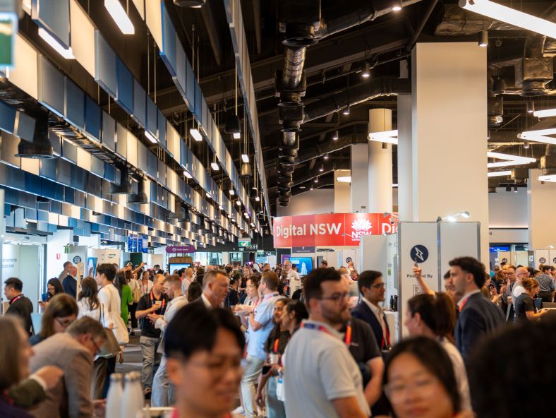 Attendees at the Digital NSW showcase event engage in conversations and explore exhibit booths in a brightly lit hall. A bright red "Digital NSW" banner stands out in the background. The space is bustling with activity, showcasing a vibrant networking environment.