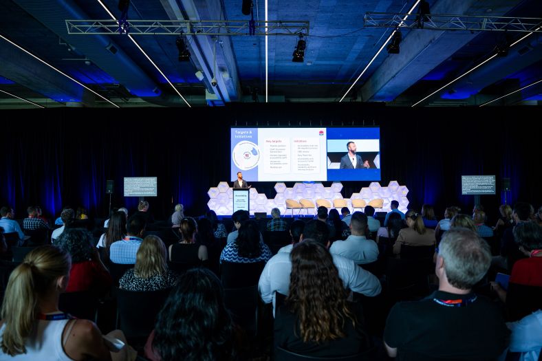 NSW Government Digital Channels Executive Director, Mark Higgs, presents to a seated audience at the Digital NSW showcase event. Modern lighting and a hexagonal backdrop frame the stage, creating a professional and dynamic atmosphere. The stage features a large screen displaying key targets and initiatives alongside the speaker. 