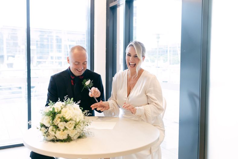 A couple sits a table by a bright window to sign their marriage certificate. They look very happy and one is laughing.