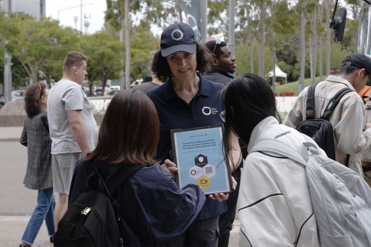 A woman working for the Sydney Olympic Park Authority holds up a clipboard with DTPR symbols for two women to look at in Sydney Olympic Park.