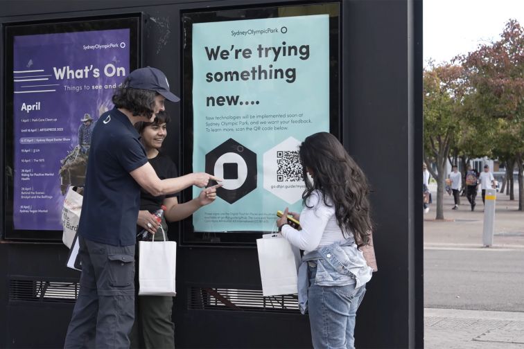 A woman working for the Sydney Olympic Park Authority points to a sign with DTPR symbols for two women to look at in Sydney Olympic Park.