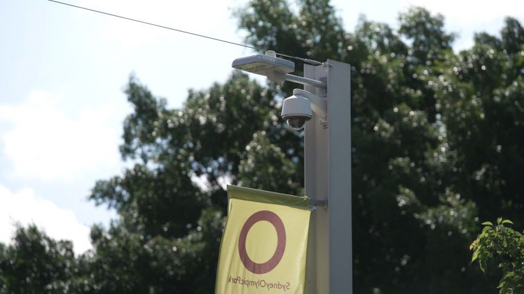 A CCTV camera is at the top of a light pole outdoors, with a Sydney Olympic Park flag flying underneath.