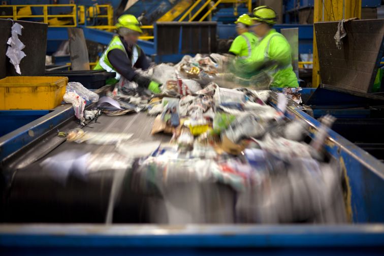 A group of people in high visibility clothing and helmets sort through waste on a recycling conveyor belt.