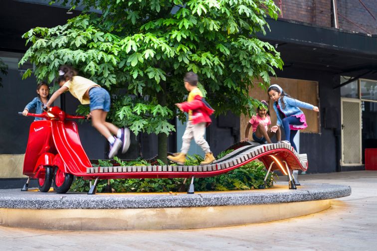 Children play on long red seat artistically designed in shape of motor scooter