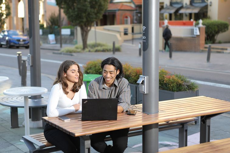 Two people are sitting outside at a ChillOUT Hub in the Georges River Council area, using the provided wi-fi and powerpoints with a laptop. Credit: Georges River Council