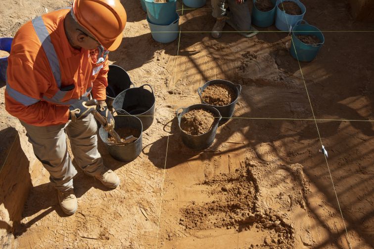 A worker wearing high-visibility clothing stoops in a dig-site, carefully operating a power tool.