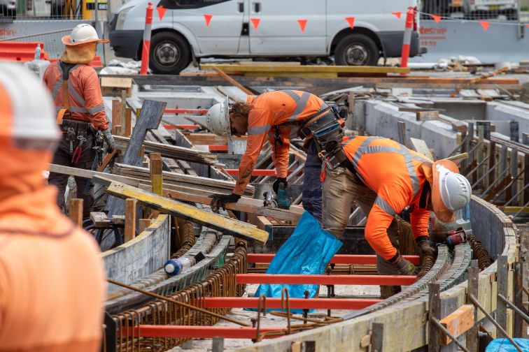 A daytime shot of construction workers wearing hi-visibility clothing and hard hats, hunched over a framework of moulding.