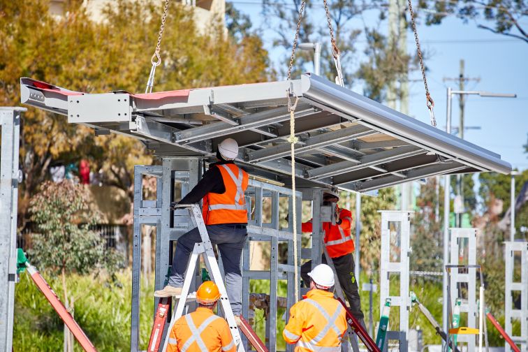 Two construction workers, wearing hi-visibility clothing, are positioned under a covering they are installing at a new light-rail station on a bright day.