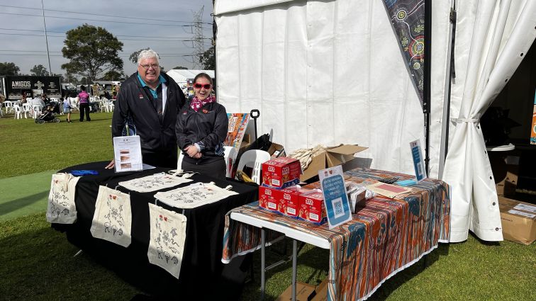 two staff members from the Aboriginal Cultural Heritage Team stand behind the ACH table at the Cooee Festival event in 2024.