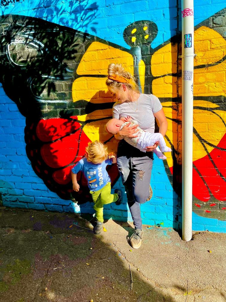 Woman breastfeeding next to an Aboriginal mural with child standing next to her