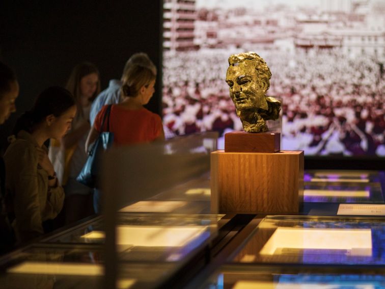 People viewing items in the exhibition gallery, a screen projects video behind them