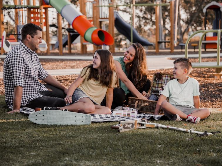 Young family sitting at a lushious green park in front of an amazing playground enjoying the sun