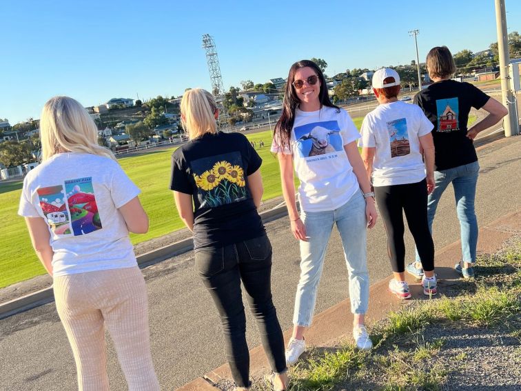 5 women in a row BH scenery behind them. Each wears a different T-Shirt featuring WNA original art.