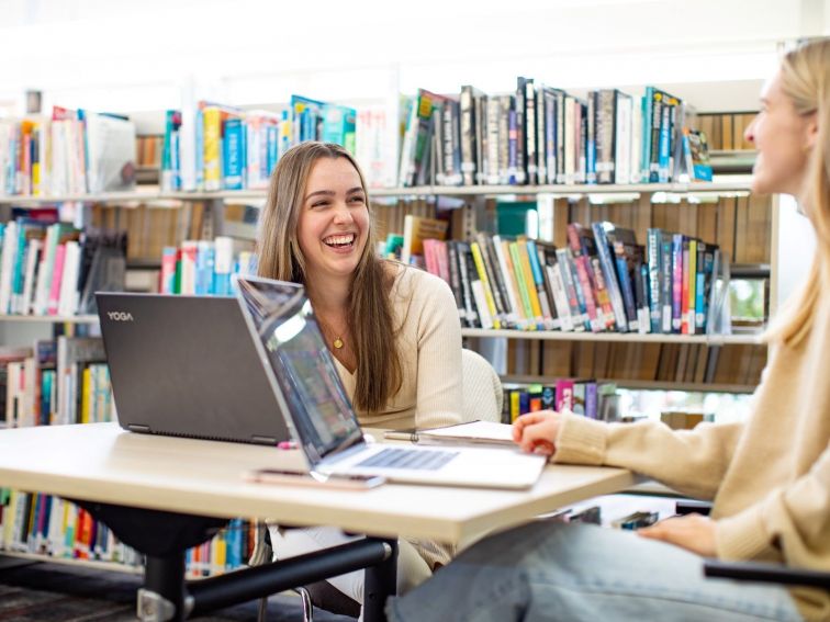 Two young women laugh while working on laptops in the library