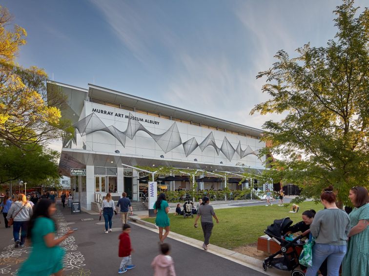 People walk in front of white rectangular art museum with cafe on terrace on the ground floor