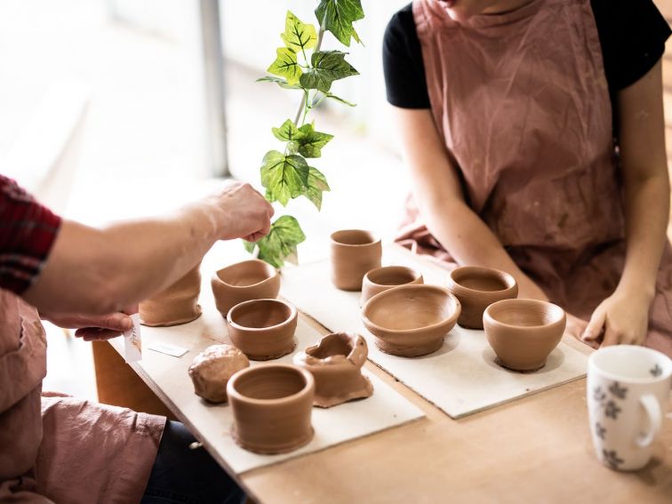 picture shows two people seated at a bench with a board of clay pots between them