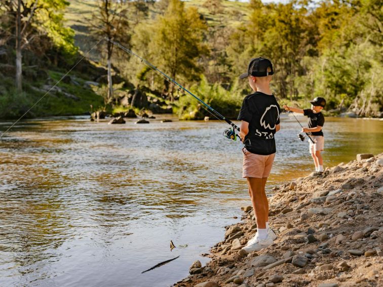 two young boys are fishing on a sandy riverbank