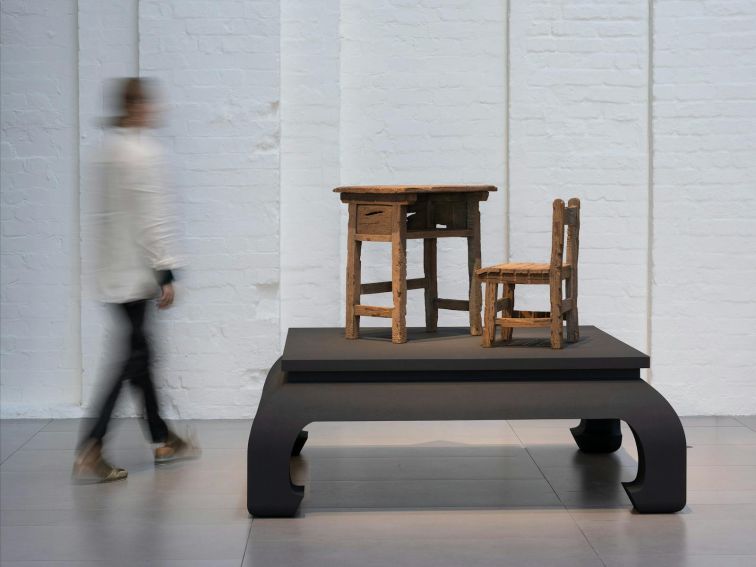 A person walking around an artwork of a table and chair made out of clay, displayed on a plinth