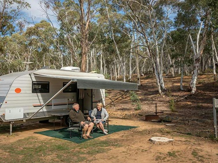 A couple sit outside their caravan in camping chairs, at Glendora campground in Hill End Historic