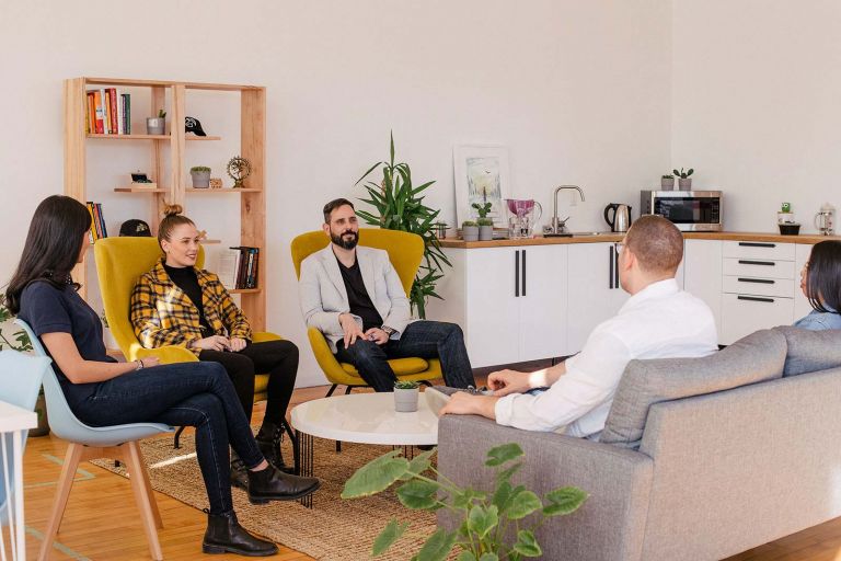 Group of five people seated around a coffee table with a kitchen and bookcase in the background