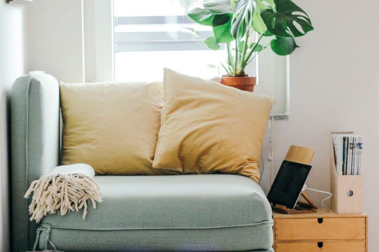 An armchair with pillows and a blanket below a window and pot plant and next to a table with a tablet and magazines