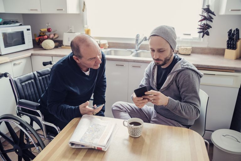 Older man in wheelchair looking at phone with younger man sitting at kitchen table