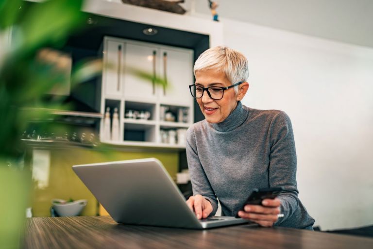 An older woman working on a laptop computer with her mobile phone doing the strata annual reporting tasks