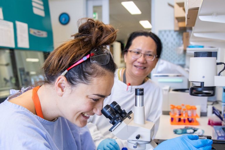 A younger woman looks through a microscope while an older woman smiles at the camera