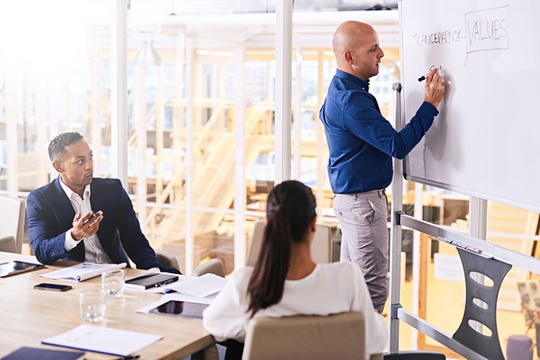 A man writing on a whiteboard in a meeting with two other people who are seated at a table