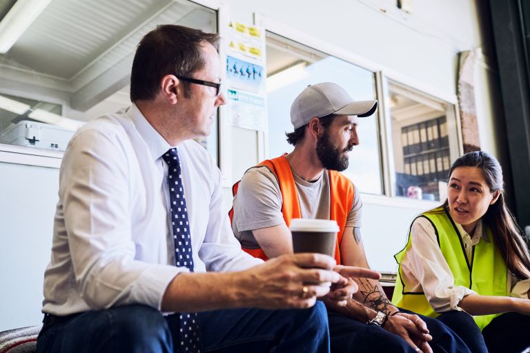 Man with white shirt, tie and cup of coffee talking to man in grey hat, shirt and orange vest and woman in yellow vest with dark long hair