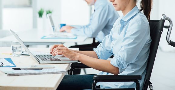 Business worker seated in wheelchair works at laptop with colleague in background