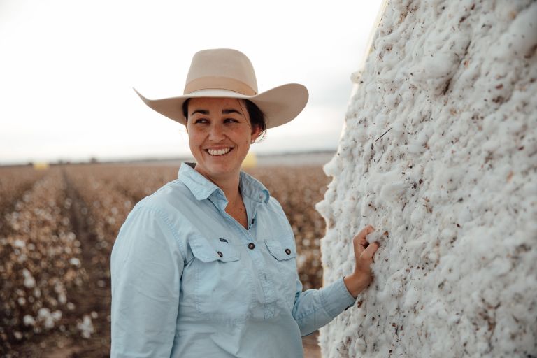 A woman is standing in a paddock up against a round bale of hay. She is wearing a light blue shirt, and cream, wide brimmed hat and is smiling.