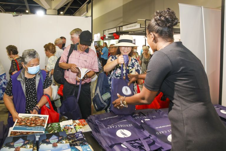 Photo of a crowd of seniors at a stall for the Seniors Card. A staff member is handing out navy blue gift bags to the smiling seniors. 