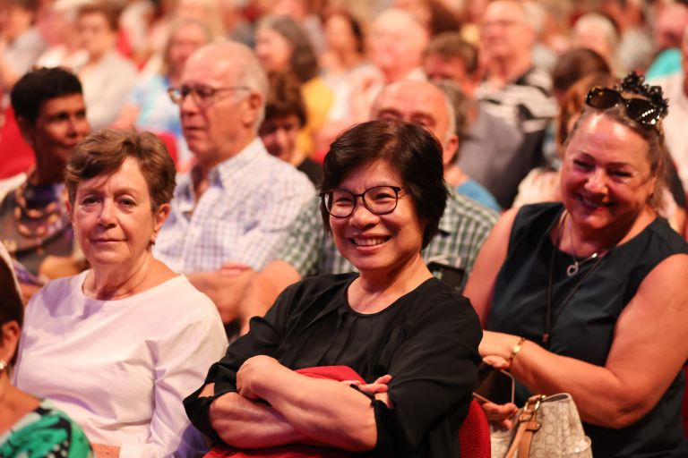 Photo focused on an Asian woman in a crowd of seated seniors. She is wear glasses, has cropped dark hair, has her arms folded and is smiling. 