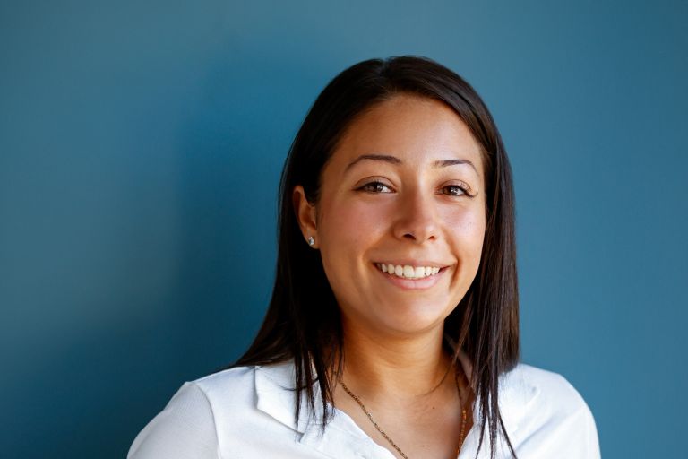 Eden is standing in front of a blue background in a white t-shirt and jeans smiling