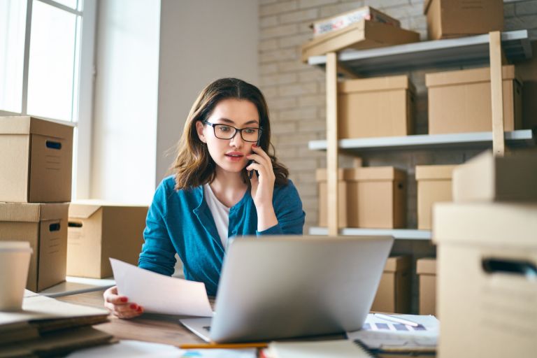 A business woman sitting in front of a laptop with cardboard boxes surrounding her.