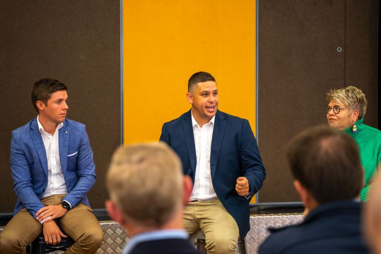 Three people sit on a panel. The man in the middle is talking. They sit in front of a yellow and brown wall.