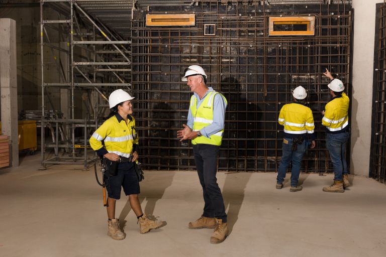 Kaitlyn talks to a colleague onsite, while other construction workers insect framework in the background.