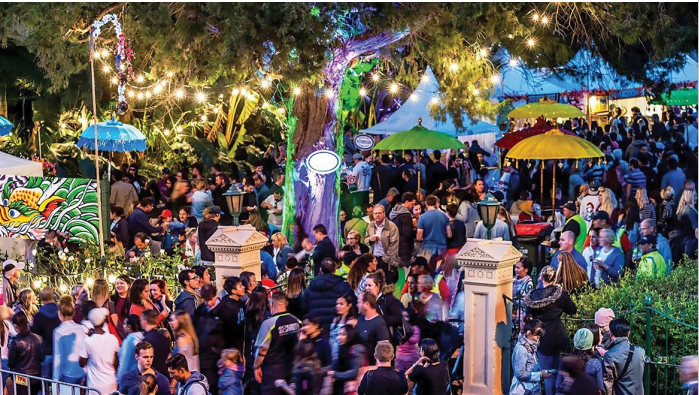 A very crowded market with tents in between trees and hanging fairy lights. 